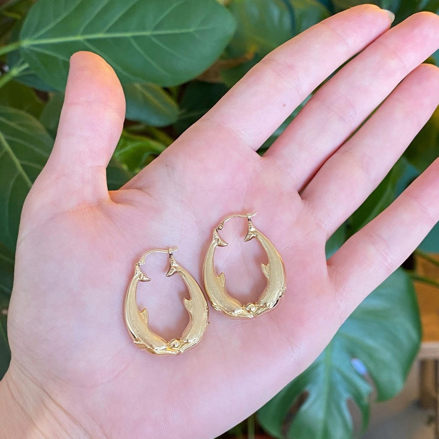 Gold hoop earrings held in a hand with a green leafy background