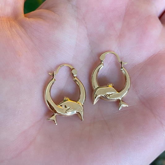 Gold dolphin earrings held in a hand with a blurred background