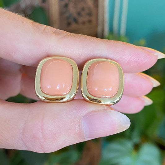 Pair of peach-colored earrings with gold accents held between fingers against a blurred green background